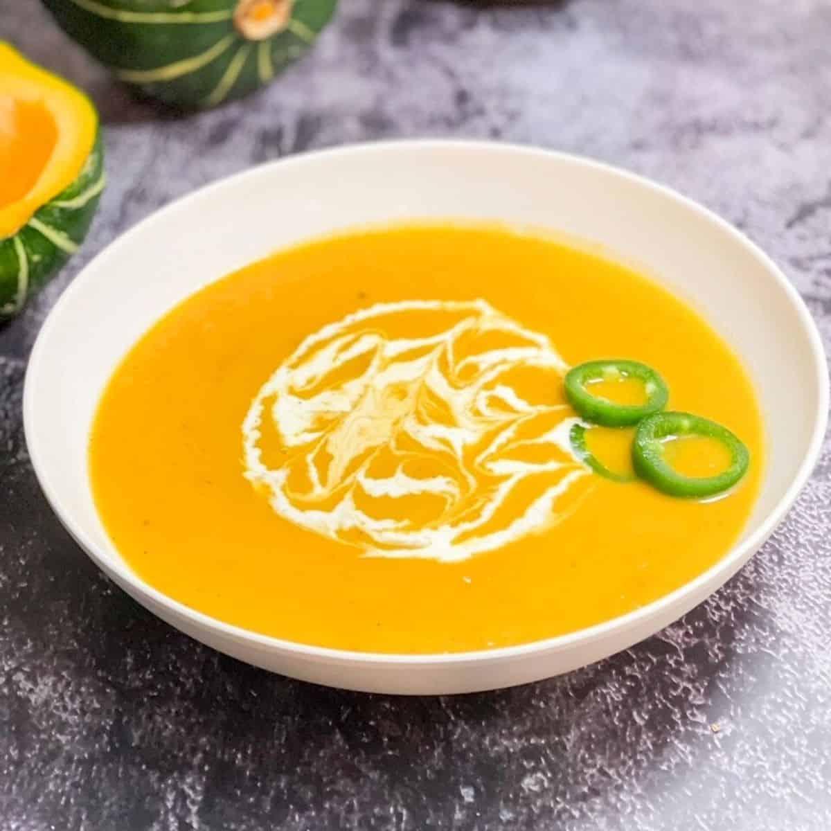 Roasted buttercup squash soup in a pale bowl with a cream swirl and green chili slices, whole squash behind