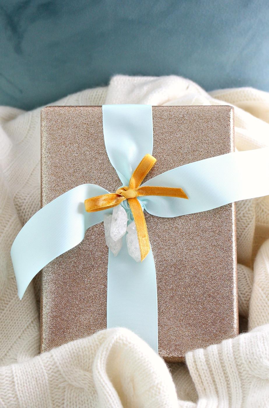 A gold glitter wrapped gift tied with pale aqua and gold velvet ribbon and topped with raw quartz crystal points