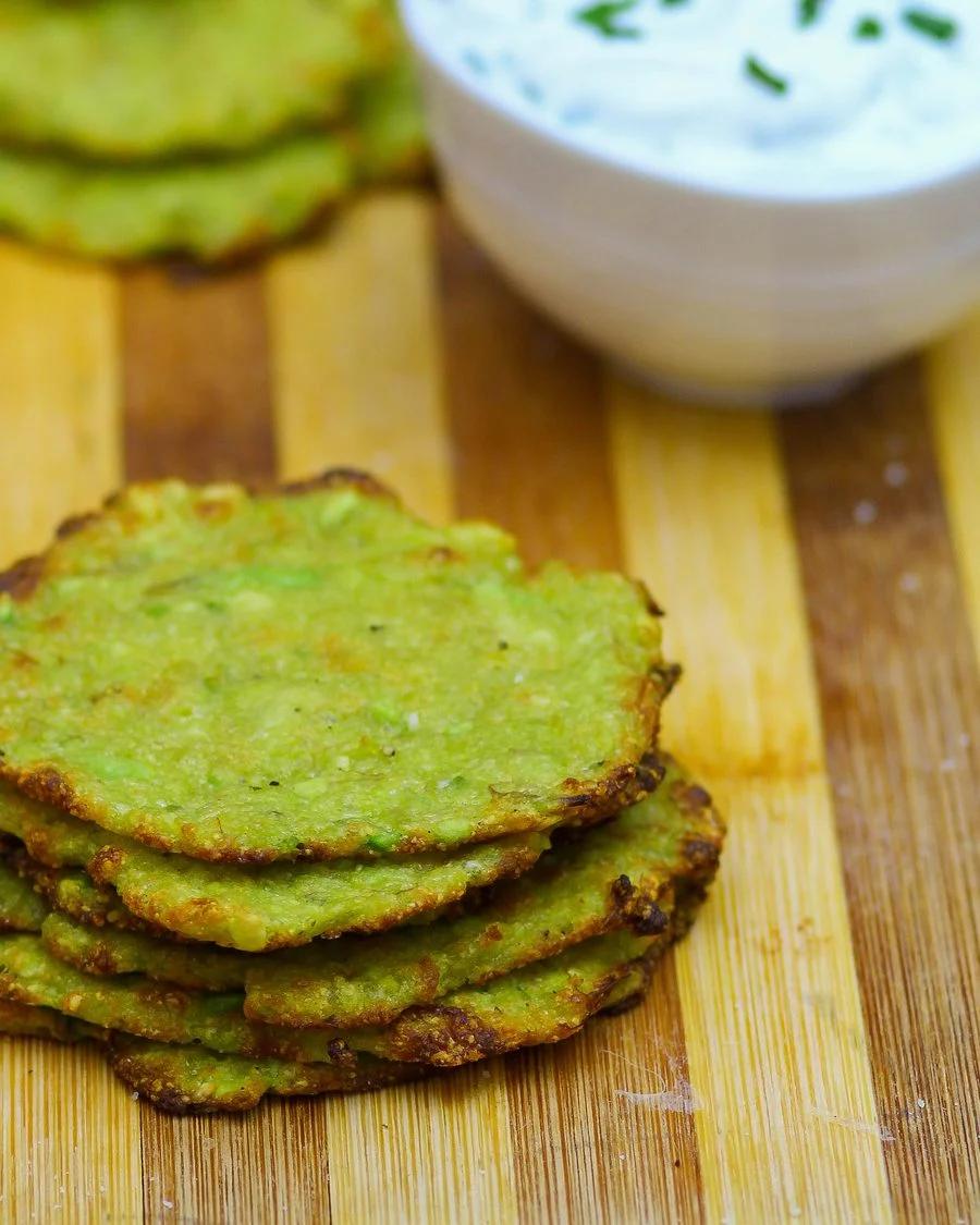 Crispy baked avocado chips stacked up on a light wooden board beside a small white bowl of ranch dip