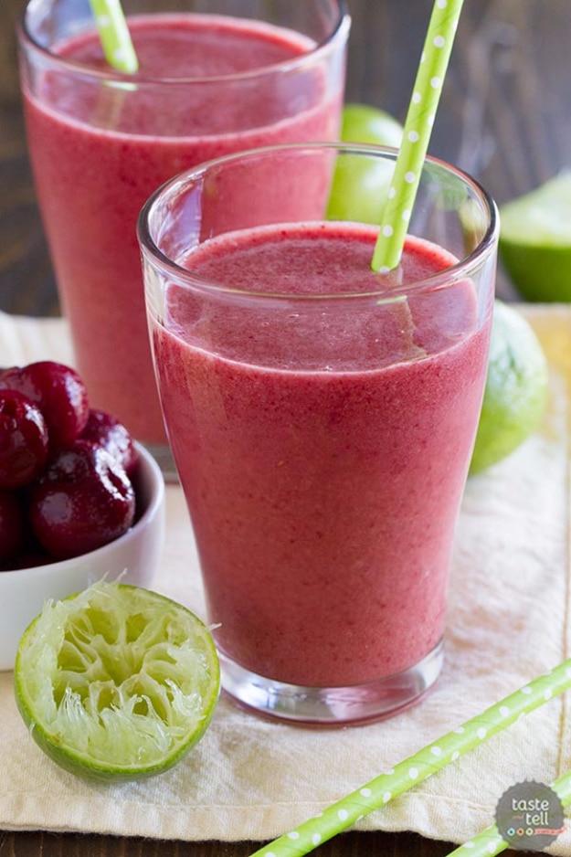 Two glasses of deep magenta cherry coconut smoothie with green straws beside fresh cherries and a squeezed lime half