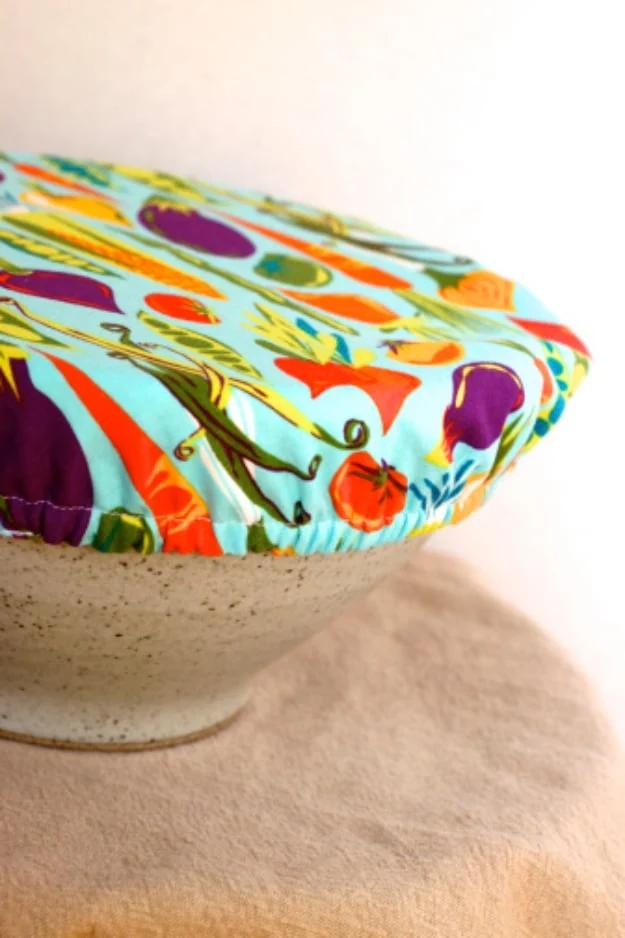 Bright vegetable print fabric bowl cover stretched over a speckled stoneware bowl against a plain pale background
