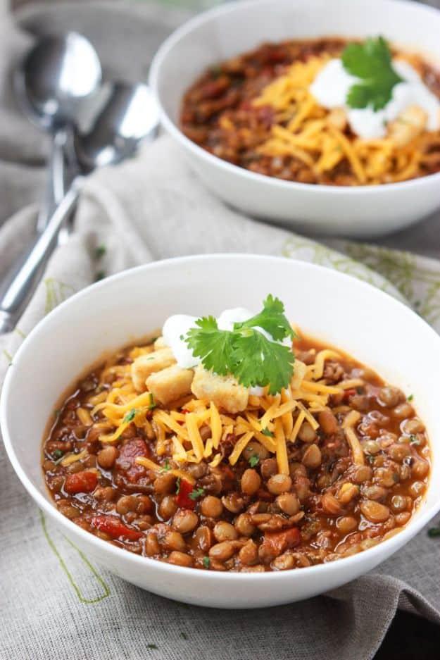 Two white bowls of lentil chili topped with sour cream, shredded cheese and fresh cilantro with spoons