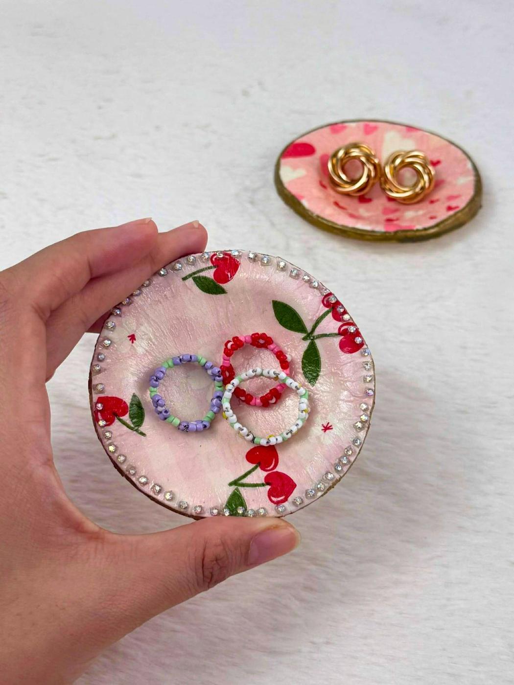 A hand holding a small clay ring dish decoupaged with cherry print paper and edged in rhinestones, a heart print dish behind