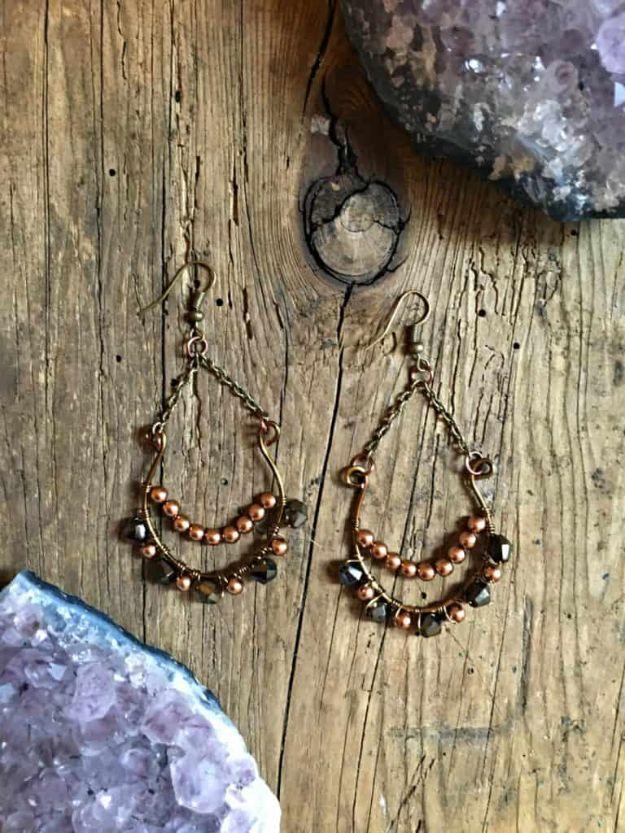Pair of brass teardrop earrings beaded with small crystals, hung on weathered wood beside amethyst geodes