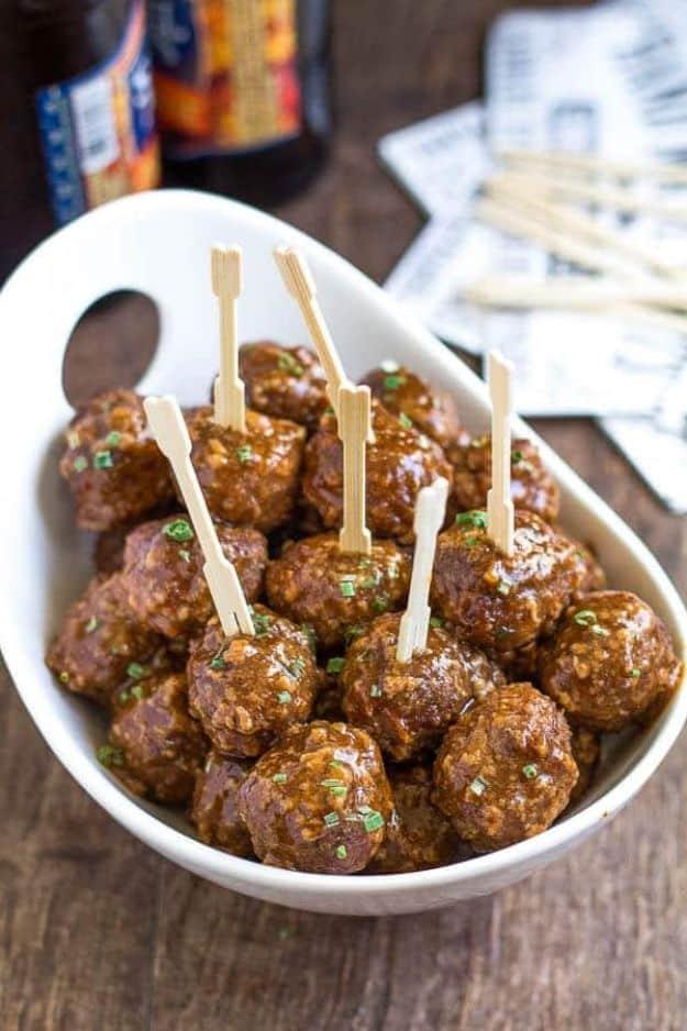 Beer BBQ meatballs glazed dark and shiny in a white bowl with wooden picks, beer bottles blurred behind