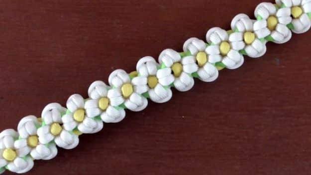 Daisy chain bracelet with white looped petals and yellow centers woven along a green cord, on dark wood