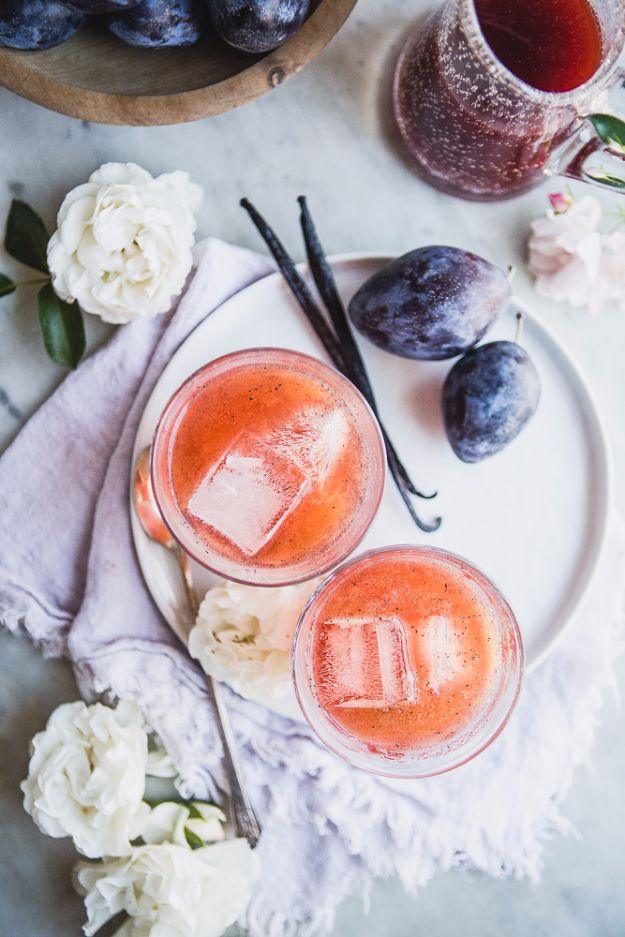 Two coupe glasses of pale pink vanilla plum shrub with club soda, surrounded by fresh plums and vanilla pods