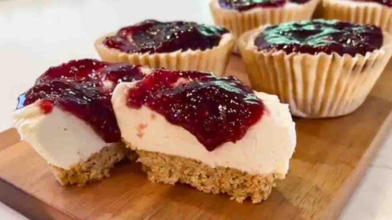 Mini no bake cheesecakes in fluted paper liners on a wooden board, topped with dark red jam, one cut clean in half