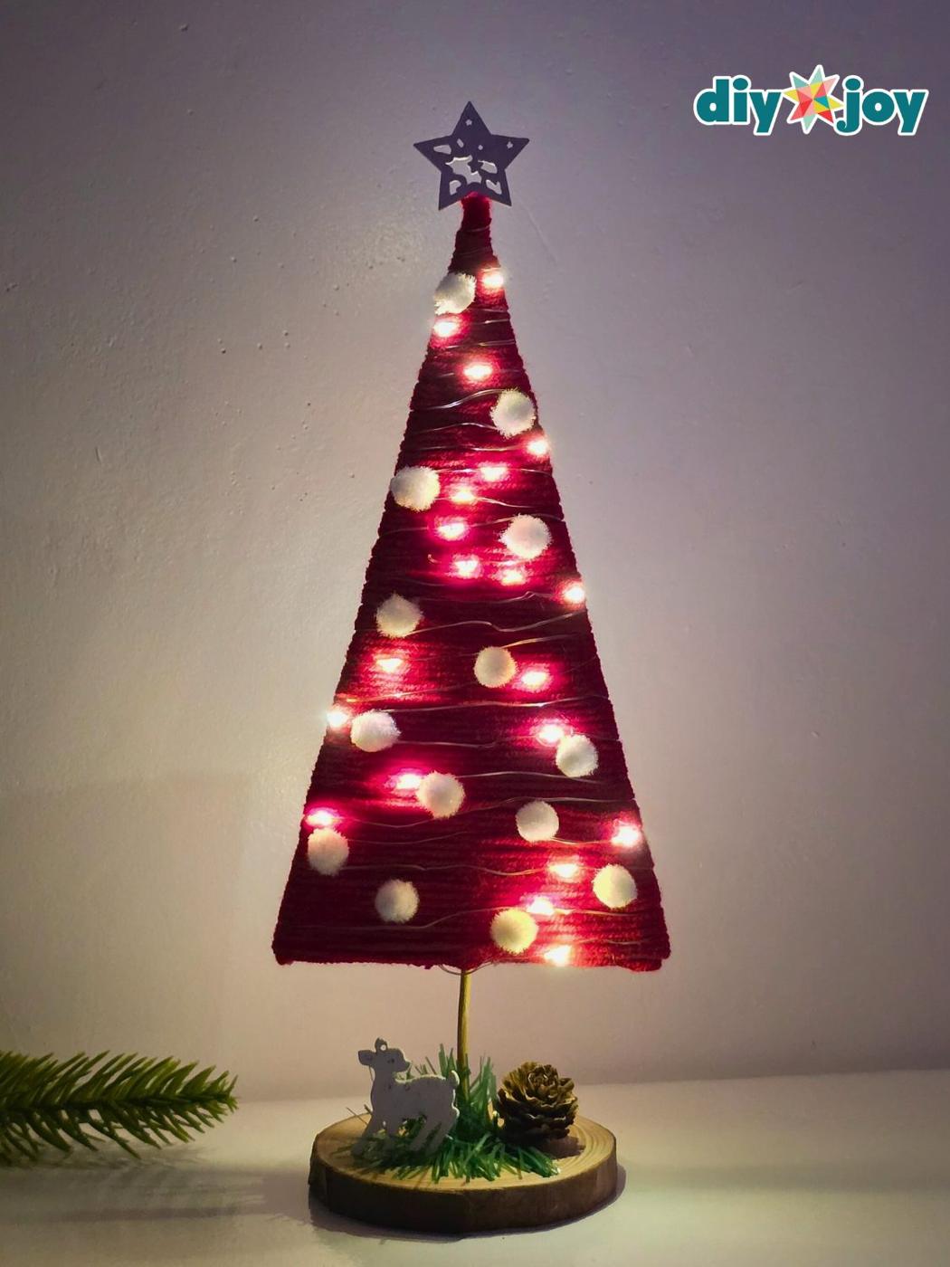 Mini Christmas tree wrapped in chunky red yarn on a cardboard triangle, lit with fairy lights and dotted with pom poms