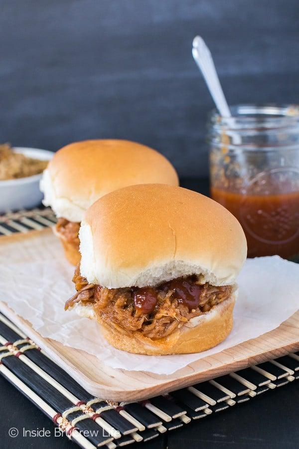 Two finished pulled-pork sandwiches on soft rolls sit on a wooden board beside a jar of barbecue sauce