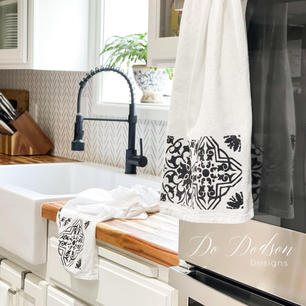 White flour sack tea towel stenciled with a black floral tile pattern, hanging from a black faucet above a farmhouse sink
