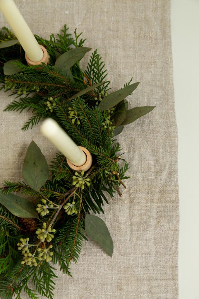A wreath of fresh evergreen and seeded eucalyptus on a linen runner with cream taper candles in small wooden holders