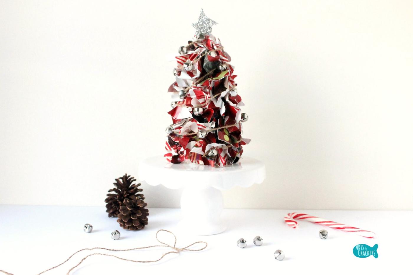 A tabletop tree of torn wrapping paper scraps with twine, silver jingle bells and a glitter star on a white cake stand