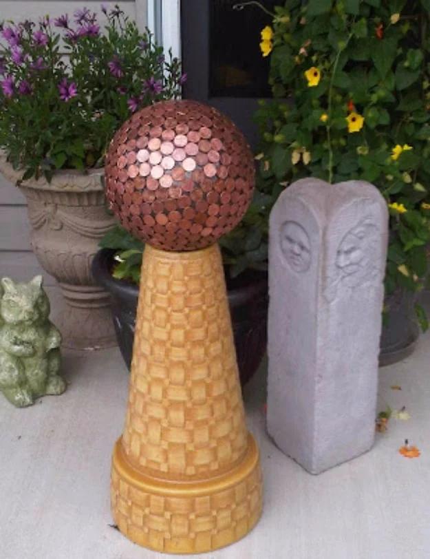Penny covered bowling ball garden gazing ball on a terracotta pedestal, surrounded by potted flowers outdoors