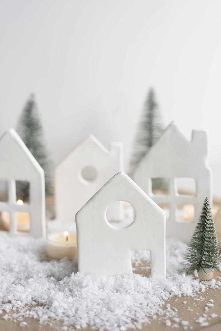 Row of small white air dry clay houses with cut out windows standing in fake snow with mini bottle brush trees