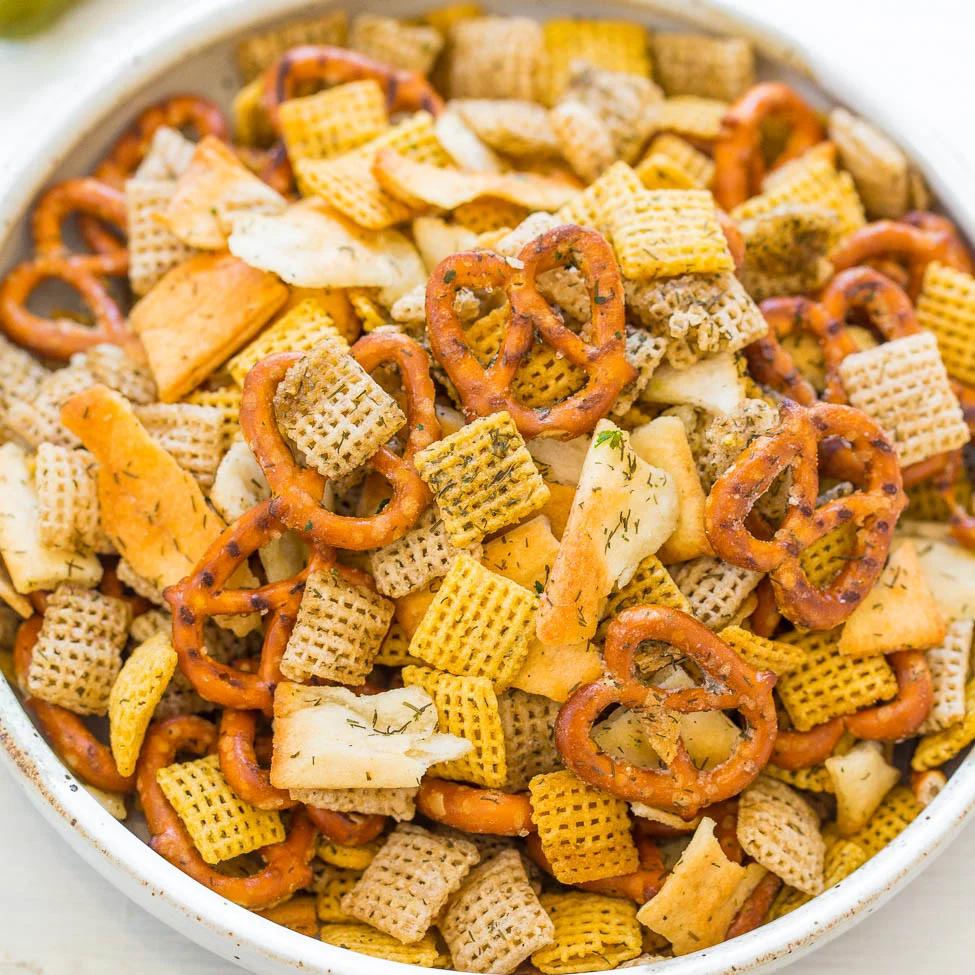 Large glass jar of dill pickle chex mix with cereal squares and pretzels, flecked with green dried dill