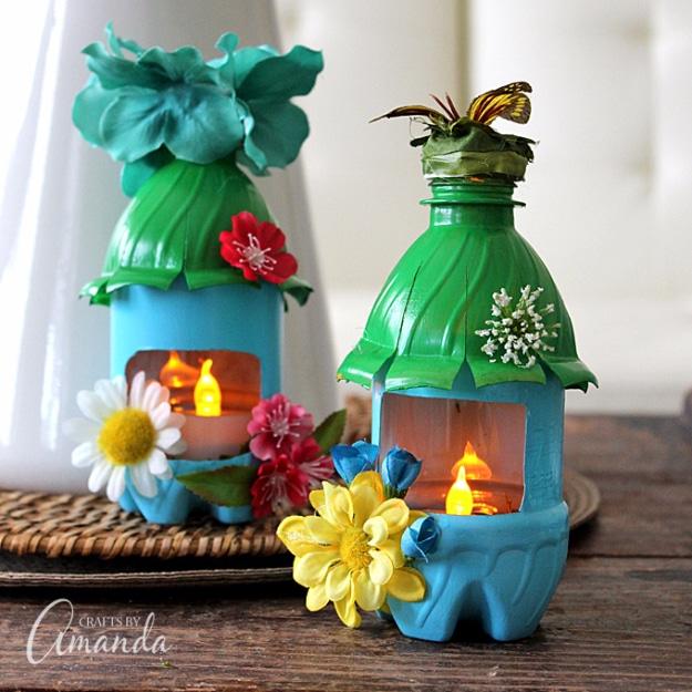 Two plastic bottle fairy houses painted blue and green with silk flower roofs and lit windows, glowing on a wooden table