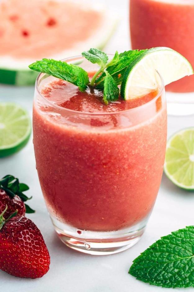 Bright red strawberry watermelon smoothie in a glass with fresh mint and a lime wedge, watermelon slices behind it