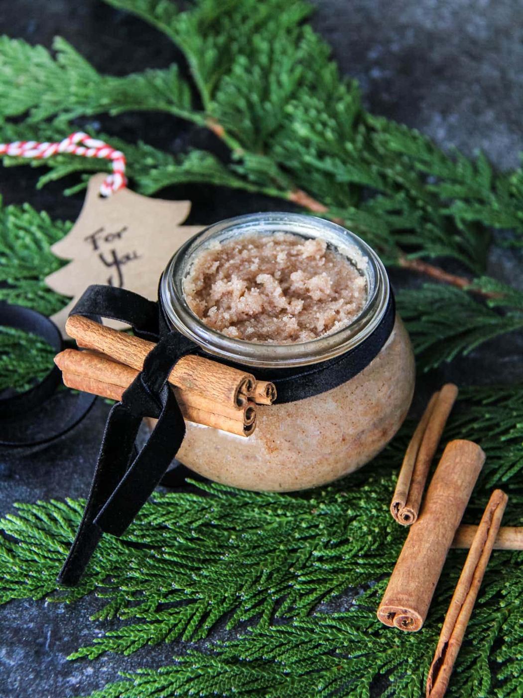 A small glass jar of cinnamon sugar scrub tied with black velvet ribbon and cinnamon sticks on a bed of fresh cedar
