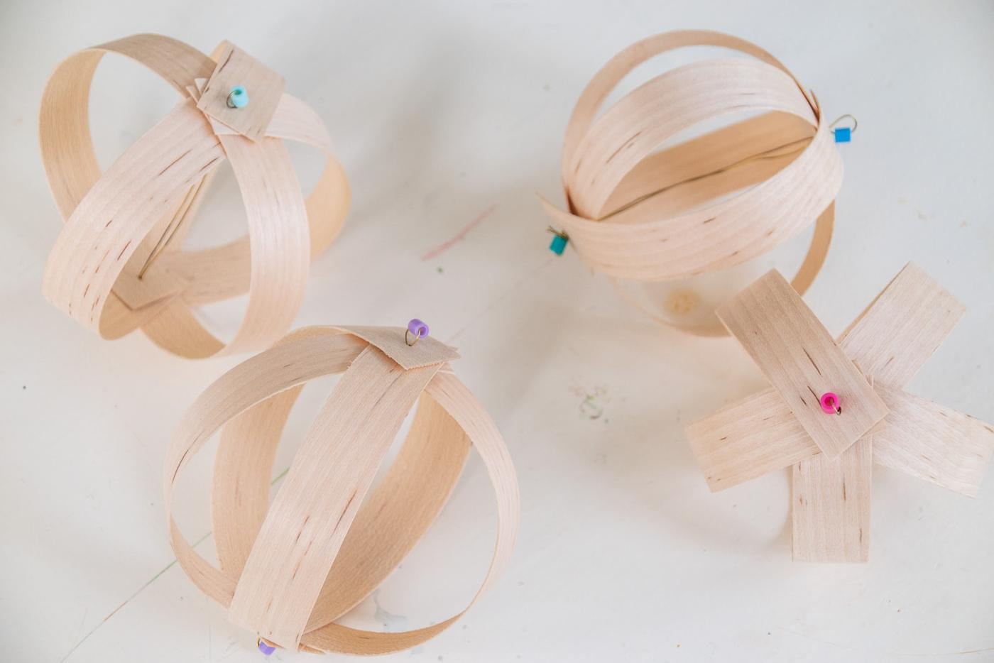 Three round ornaments of curved pale wood veneer edge banding strips with tiny colored beads on top, on a white table