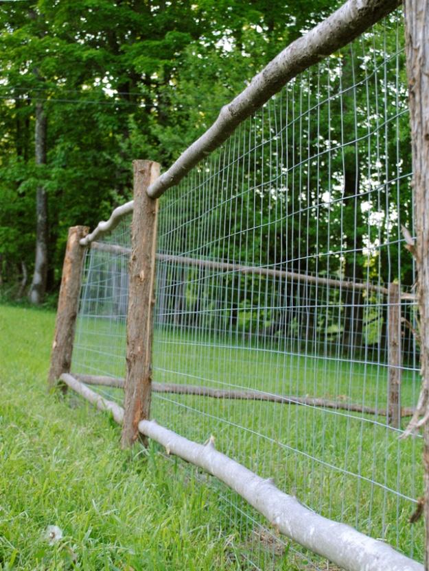 Rustic garden fence, rough sapling posts and rails with wire mesh behind them running along a green lawn beside trees