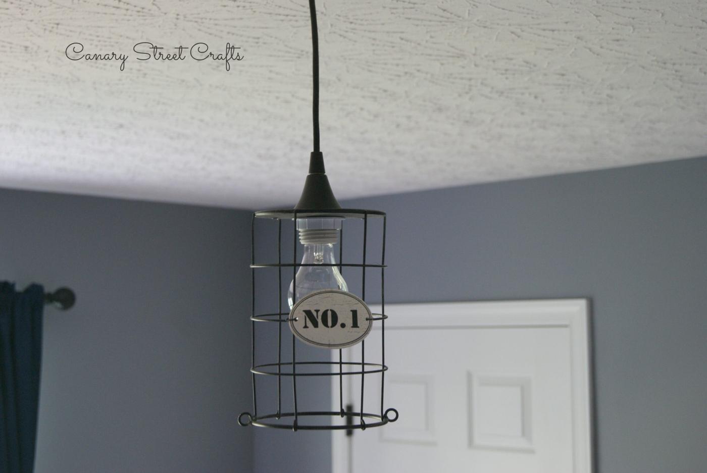 Black wire cage pendant light with an Edison bulb and a No 1 tag, hanging from a white ceiling by a grey wall