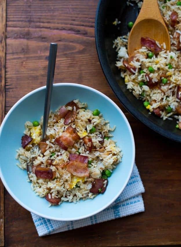 Bacon fried rice in a blue bowl with peas and bacon pieces, a dark skillet of more rice behind on wood