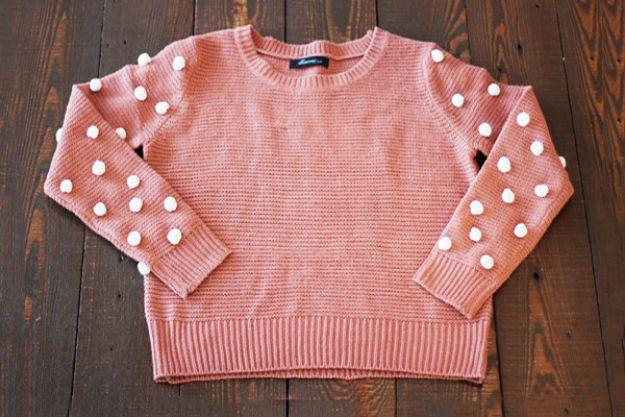 DIY clothes for winter, a pink knit sweater laid flat on dark wood with white pom poms sewn along both sleeves