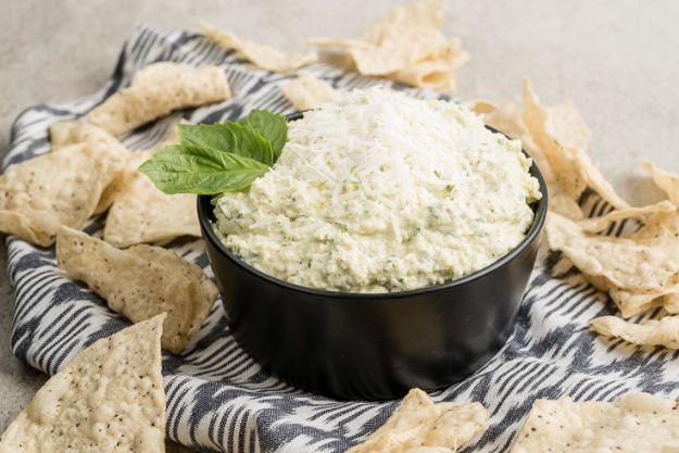 Creamy goat cheese and artichoke dip in a black bowl topped with a basil leaf, surrounded by tortilla chips