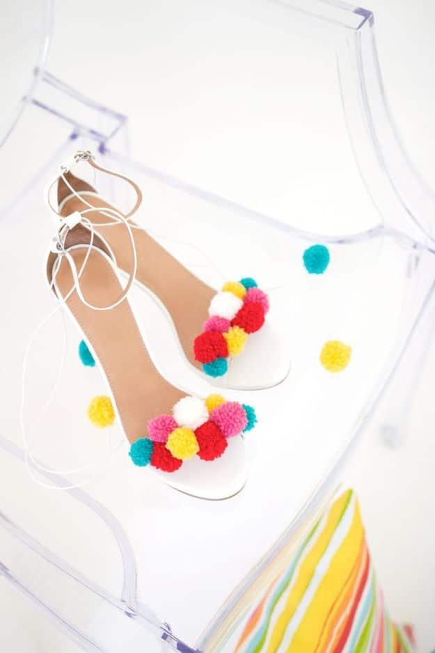 White ankle strap sandals covered with rows of pink, yellow and teal yarn pom poms, on a clear acrylic chair