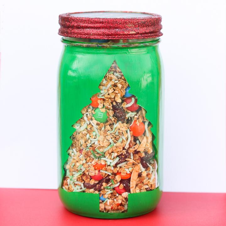 A quart mason jar painted green with a Christmas tree window showing granola and red and green candies, with a glitter lid