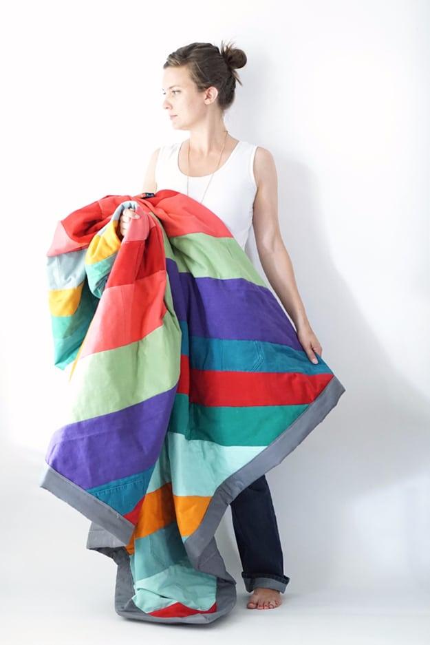Woman holding up a large rainbow striped patchwork quilt with gray binding against a plain white wall
