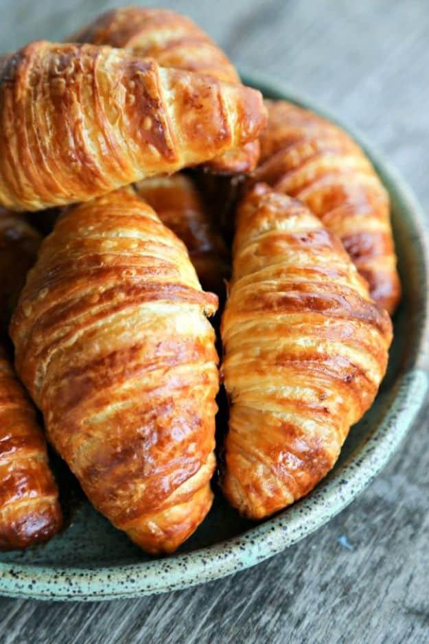 Homemade croissants baked deep golden brown with flaky layered ends, piled in a green ceramic bowl on a grey table