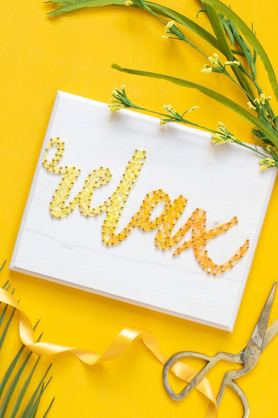 White wood board with the word relax in yellow ombre string art on gold nails, on a yellow background with scissors