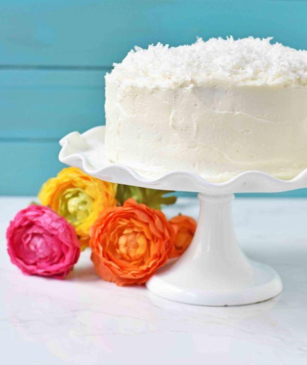 Tall white coconut cake covered in shredded coconut on a ruffled cake stand against a turquoise wall