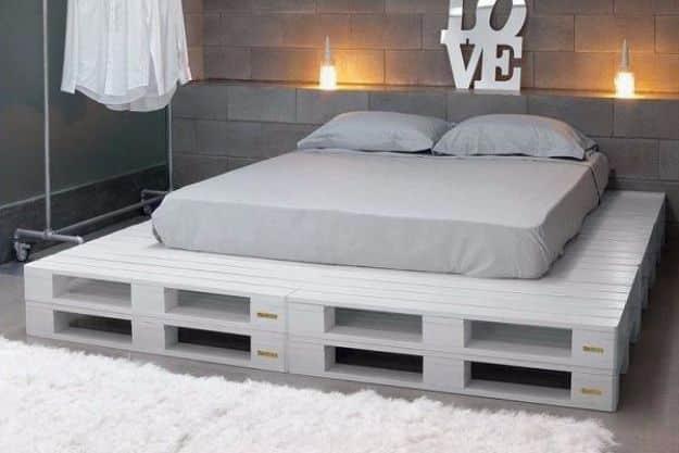 Photograph of a white painted pallet platform bed with gray bedding against a gray block wall with candles
