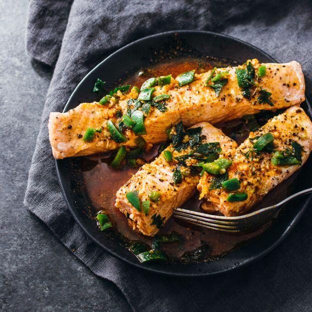 Three salmon fillets topped with chopped green chili and cilantro on a dark plate with sauce and a fork