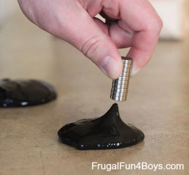 Make magnetic slime, an adult hand holding a stack of silver disc magnets above black slime pulled up into a tall sharp cone