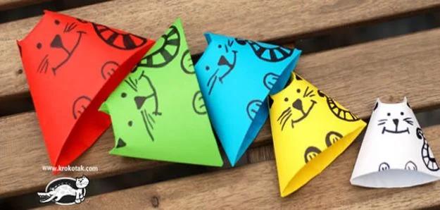 Five folded paper cats in red, green, blue, yellow and white with drawn faces and whiskers, in a row on a wooden bench