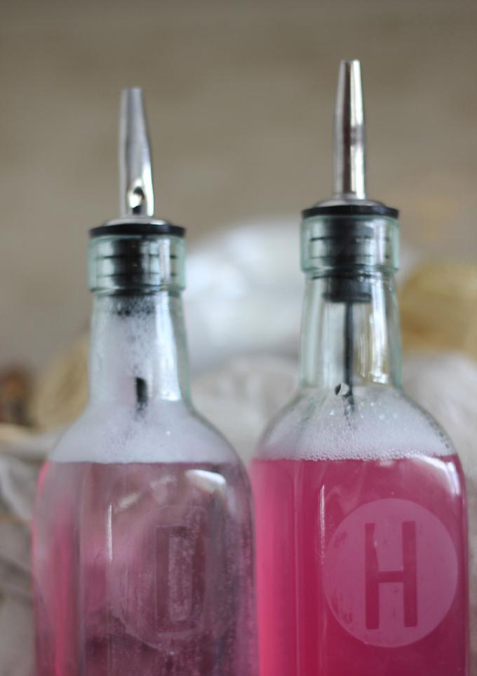 Two glass oil dispenser bottles of pink soap etched with the letters H and D and fitted with metal pour spouts