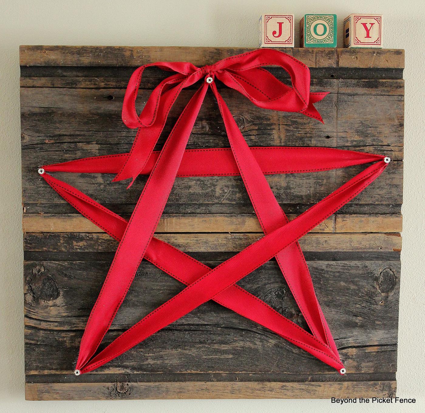 Red ribbon star stretched between five screws on a square of weathered gray barn wood, tied with a bow at the top