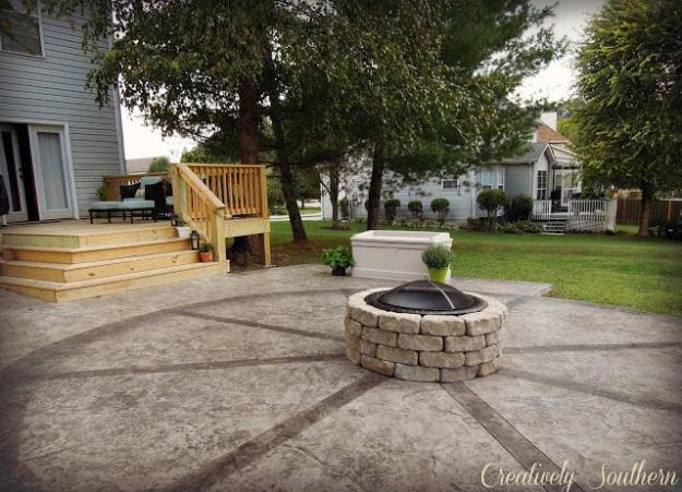 Round stacked stone fire pit with a black metal bowl set on a stamped concrete patio beside a wooden deck