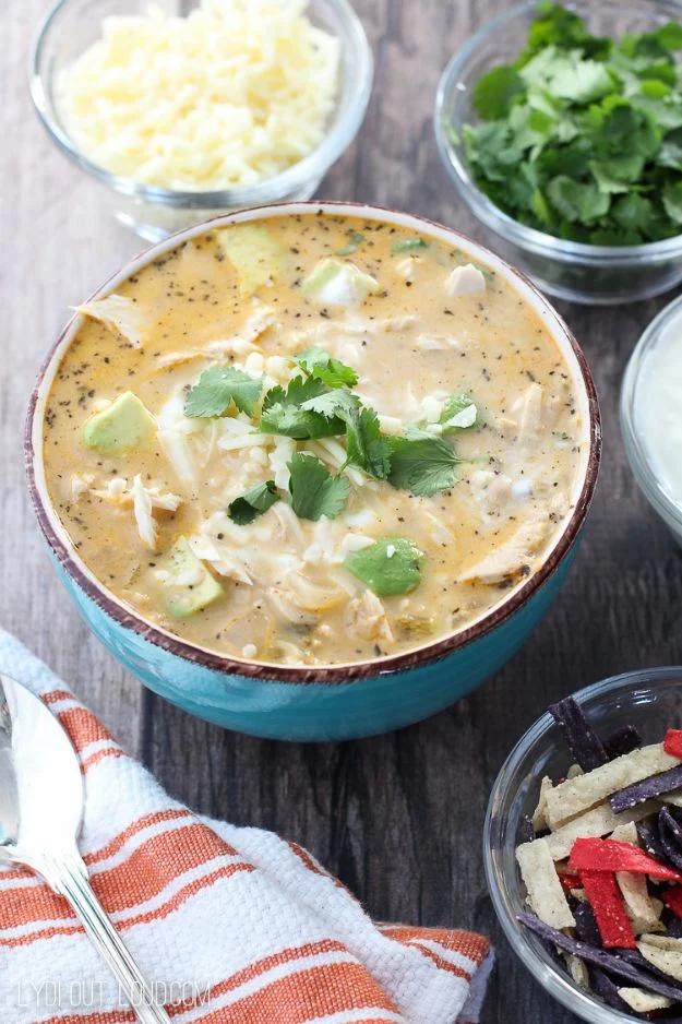 Queso white chicken chili in a teal bowl with cilantro and small bowls of cheese and tortilla strips