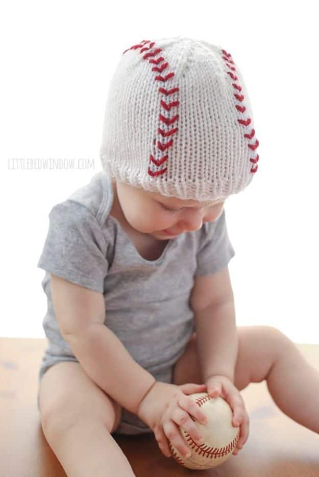 White knitted baby hat with red baseball seam stitching, worn by a baby holding a real baseball in both hands