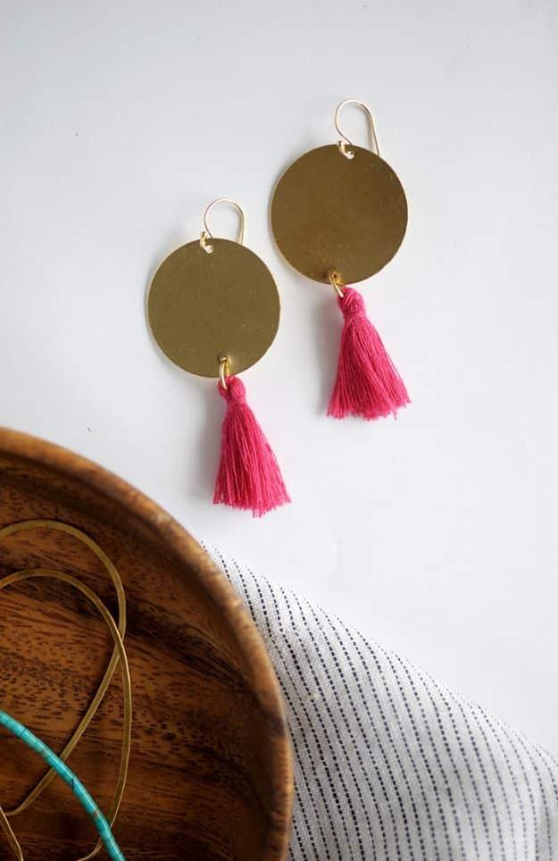 DIY statement earrings, large brass discs with hot pink tassels hanging below, beside a wooden bowl and striped cloth