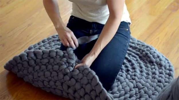 Woman kneeling on a wood floor finger crocheting a thick grey merino wool roving DIY rug in a spiral