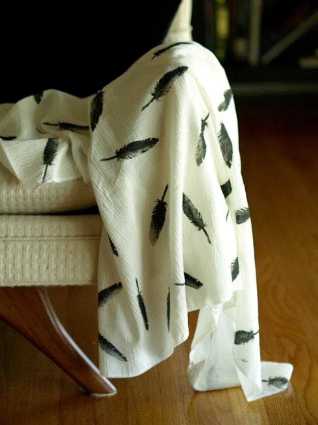 Cream cotton gauze baby swaddle stamped with scattered dark feathers, draped over a pale wooden bench