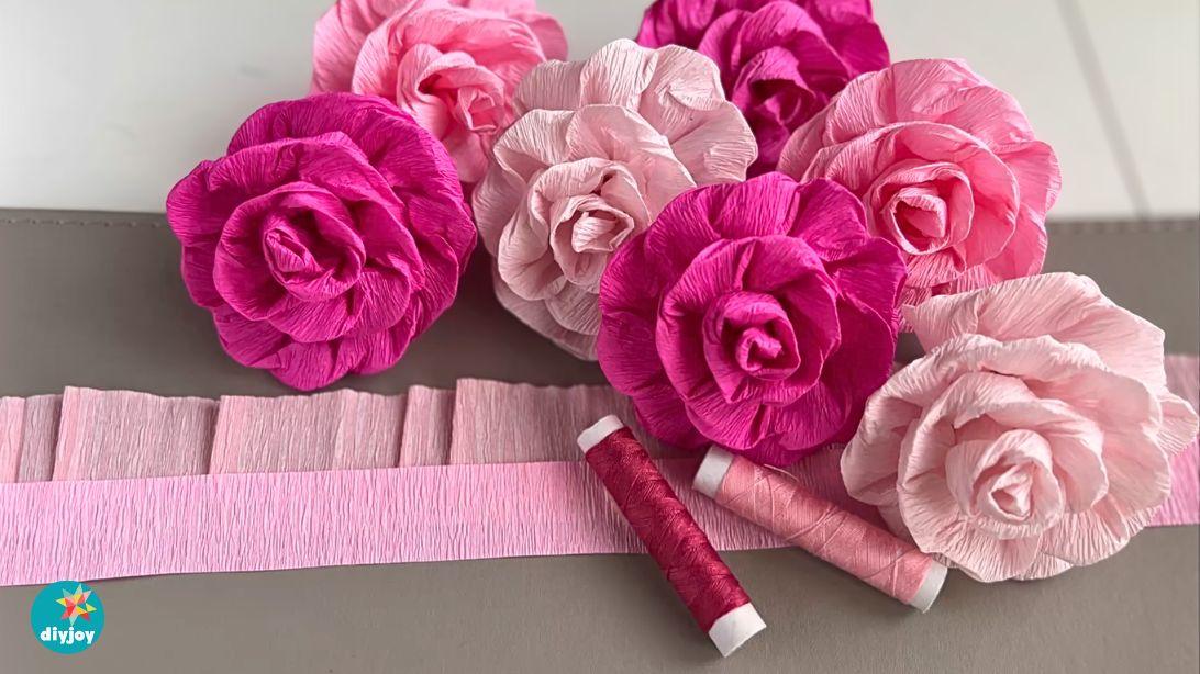 Crepe paper flowers, pink and magenta crepe paper roses beside long fringed crepe strips and rolls on a gray surface