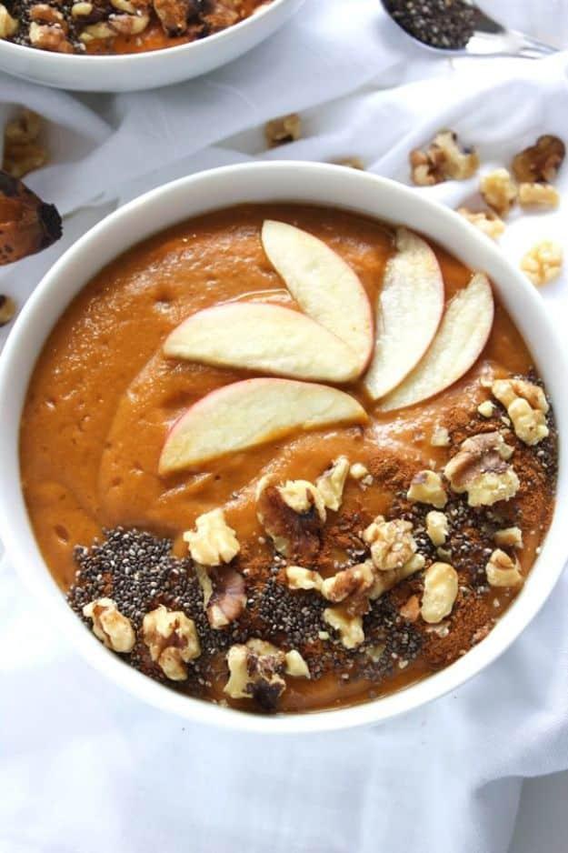 White bowl of a thick orange smoothie topped with apple slices, walnuts and chia seeds on a white cloth