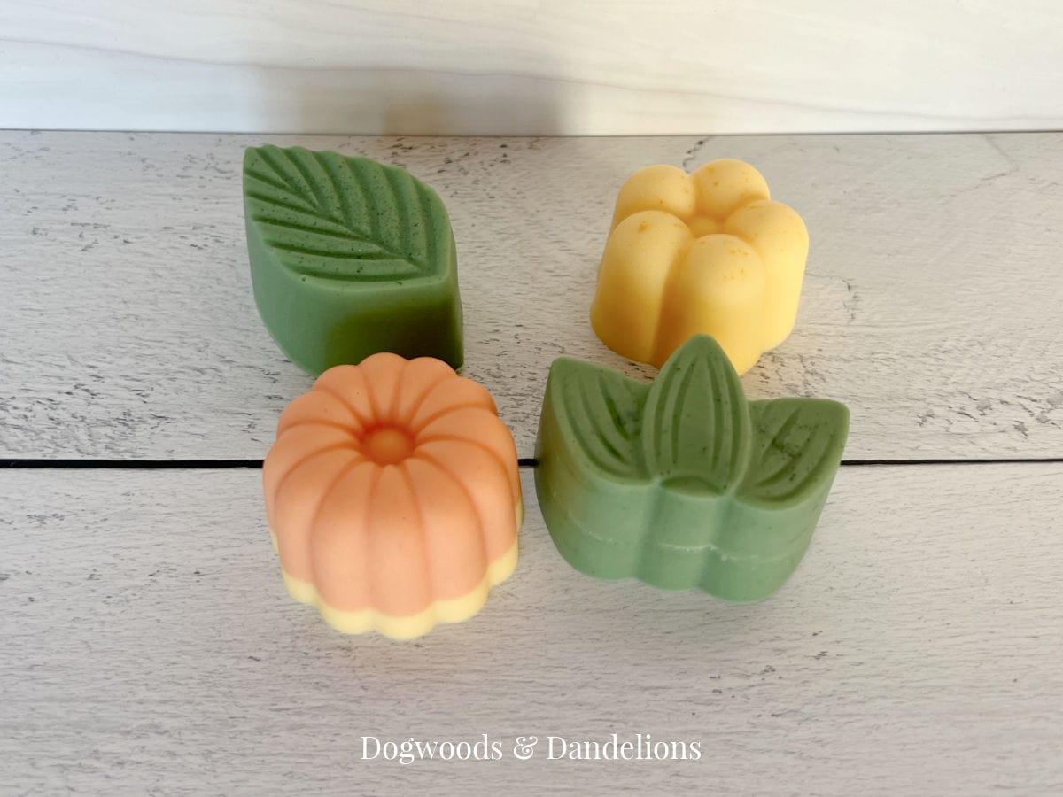 Four molded decorative soap bars in green, yellow, orange and cream shaped like leaves and flowers on white wood
